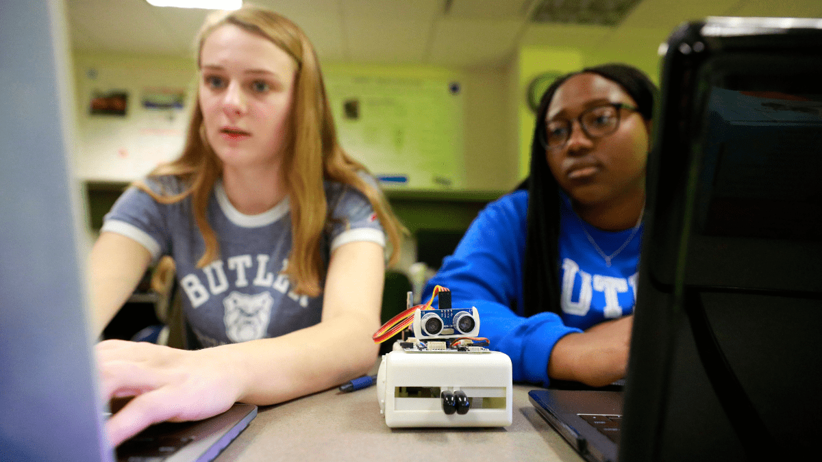 Two female students work with computers and a robot