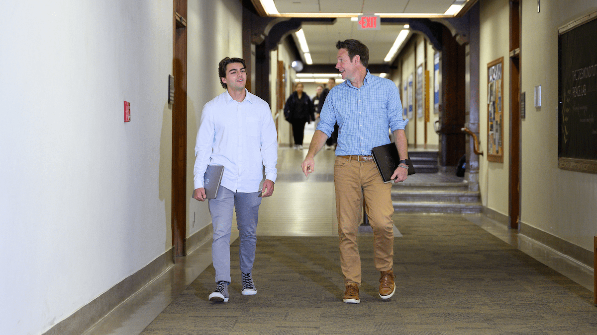 Two men walking in a hallway, one in a white shirt and gray pants, the other in a blue shirt and tan pants. both carrying a laptop computer.