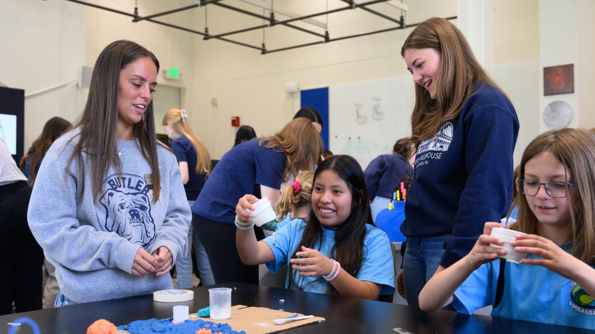Two female Butler students in the Elementary and Middle Secondary programs, wearing Butler crewnecks, and standing at a table with two young girls wearing light blue t-shirts, from two local elementary schools, engaging with moon rocks and meteorite samples from NASA.